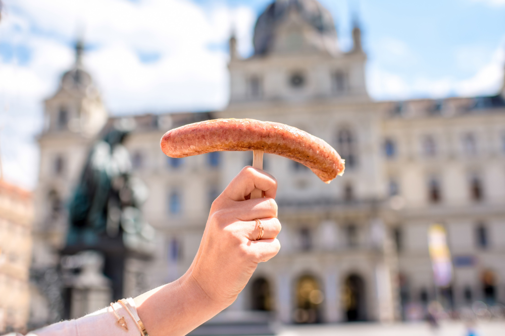 Historic city square, grilled sausage on stick foreground, statue and buildings blurred, midday sun, editorial travel photo, no people.
