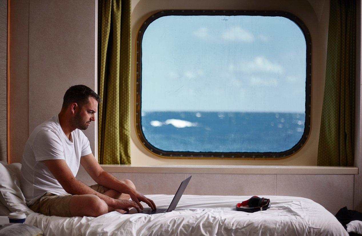 Ocean-view bedroom, man on bed with laptop, window framing sky and sea, daytime natural light, editorial travel photo, one person.