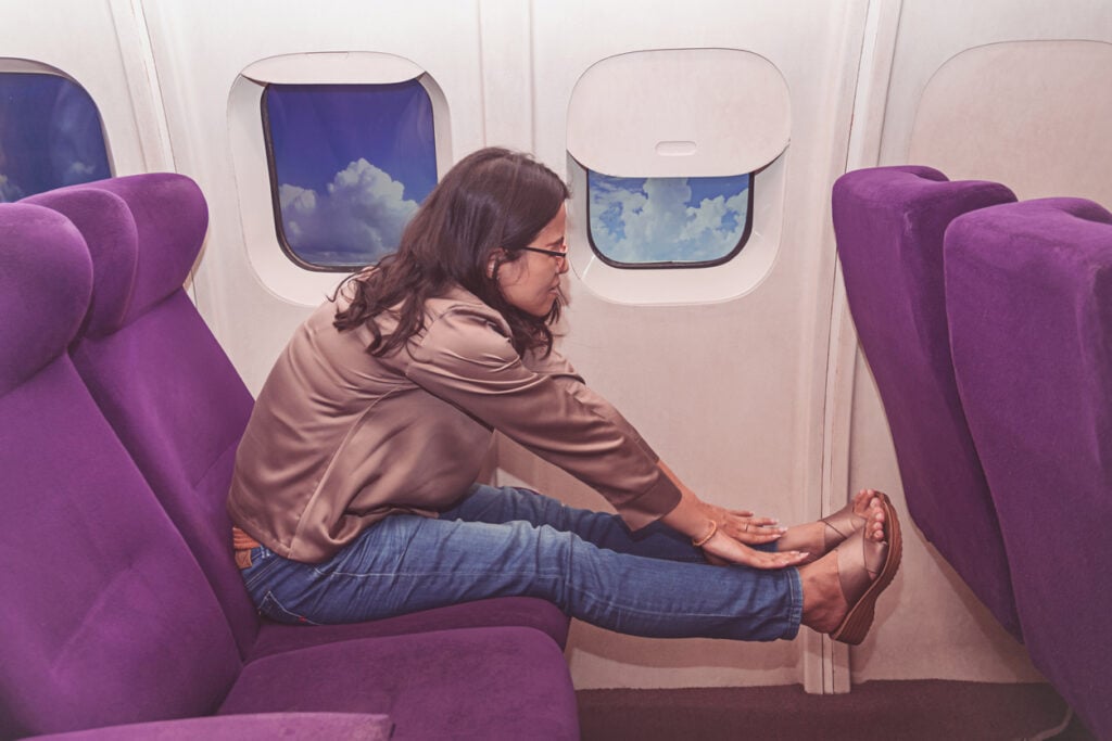 Aircraft cabin, woman stretching in purple seats, side view with visible blue sky through windows, daytime, editorial travel photo, one person.