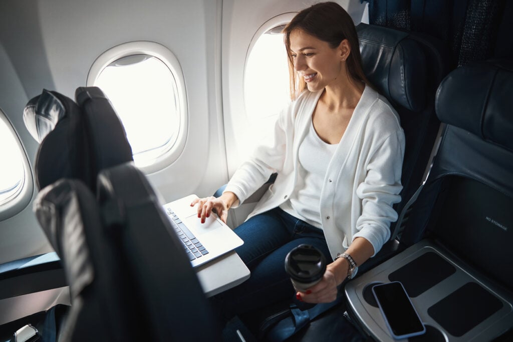 Airplane cabin, woman with laptop and coffee, window seat composition, daylight, editorial travel photo, one person.