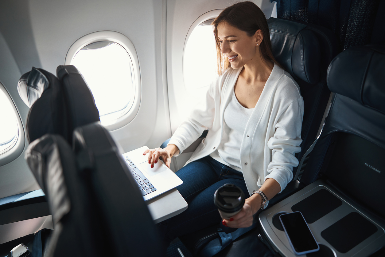 Airplane cabin, woman with laptop and coffee, window seat composition, daylight, editorial travel photo, one person.