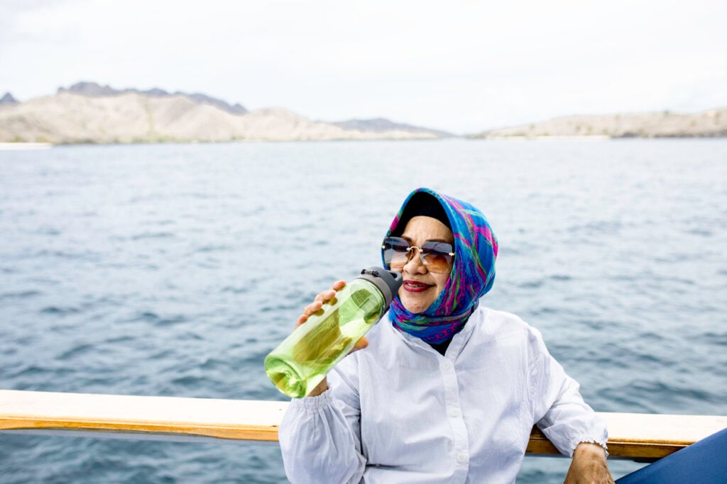 Boat deck, older woman in bright headscarf with sunglasses, drinking water bottle, afternoon sun, editorial travel photo, people.