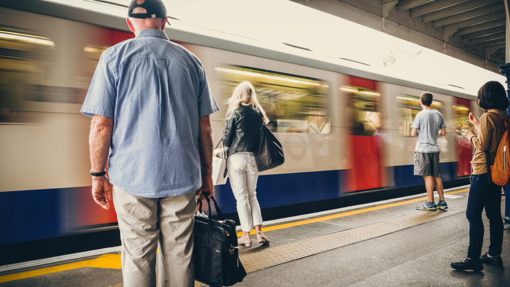 Train platform, subway train in motion blur, partial roof overhead, daylight scene, editorial travel photo, several people.