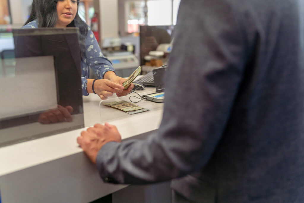 Bank interior, cash passing between teller and suited client, close-up at counter glass, daytime lighting, documentary style, two people.
