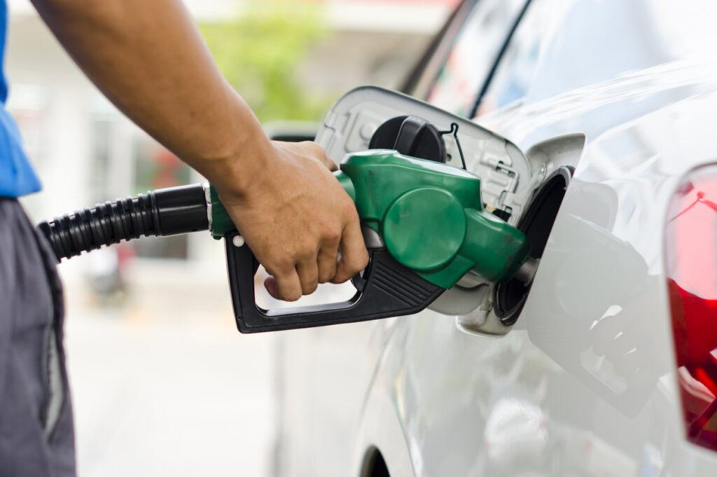 Gas station, silver car refueling, close-up on green fuel nozzle, daytime lighting, editorial travel photo, person visible.