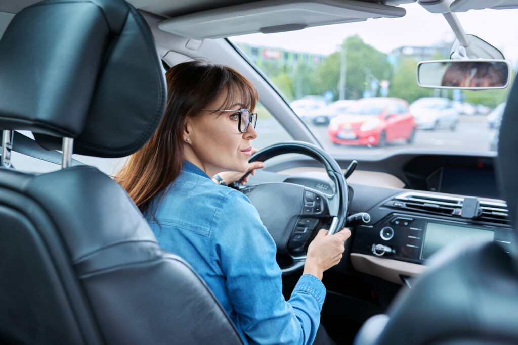Parking lot, woman reversing, over-shoulder view from front seat, daylight, documentary style, one person visible.