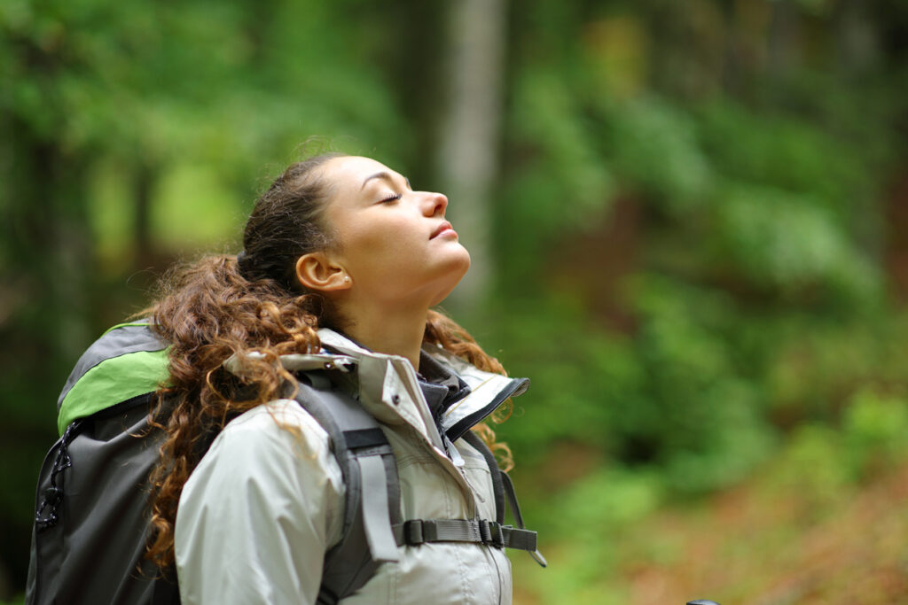 Lush forest, woman with curly hair in gray jacket, mid-ground centered, daylight, editorial travel photo, one person.