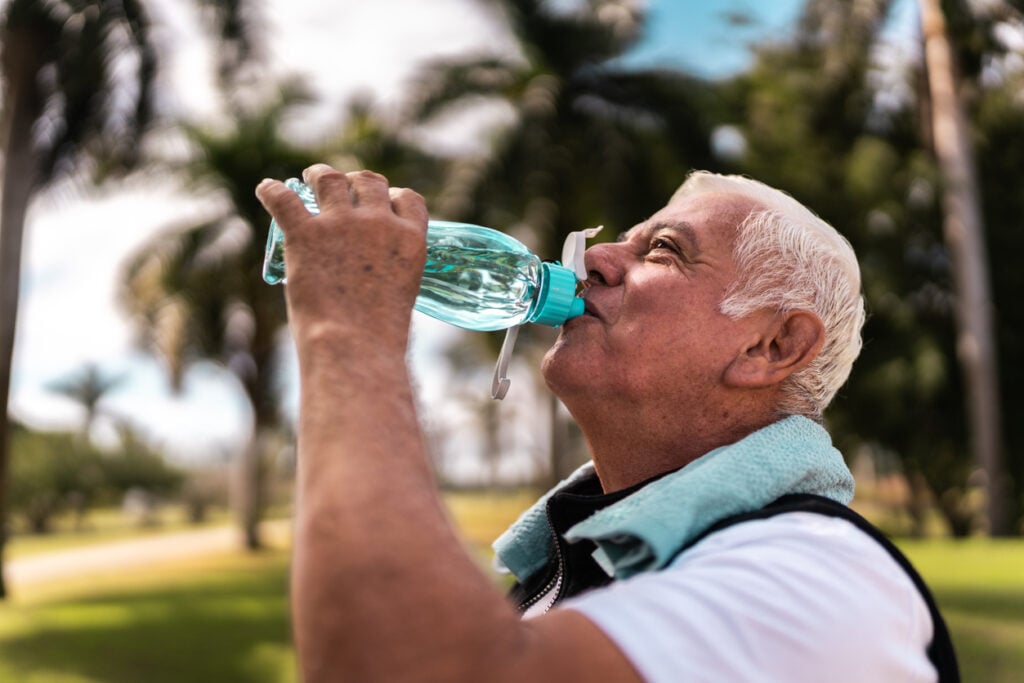 Sunny outdoor park, man with white hair drinking from bottle, towel on neck, palm trees, daytime light, editorial travel photo, people.