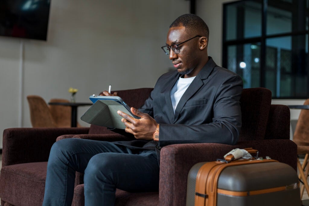 Modern lounge, businessman with tablet and stylus, seated near suitcase, bright natural light, editorial travel photo, one person.