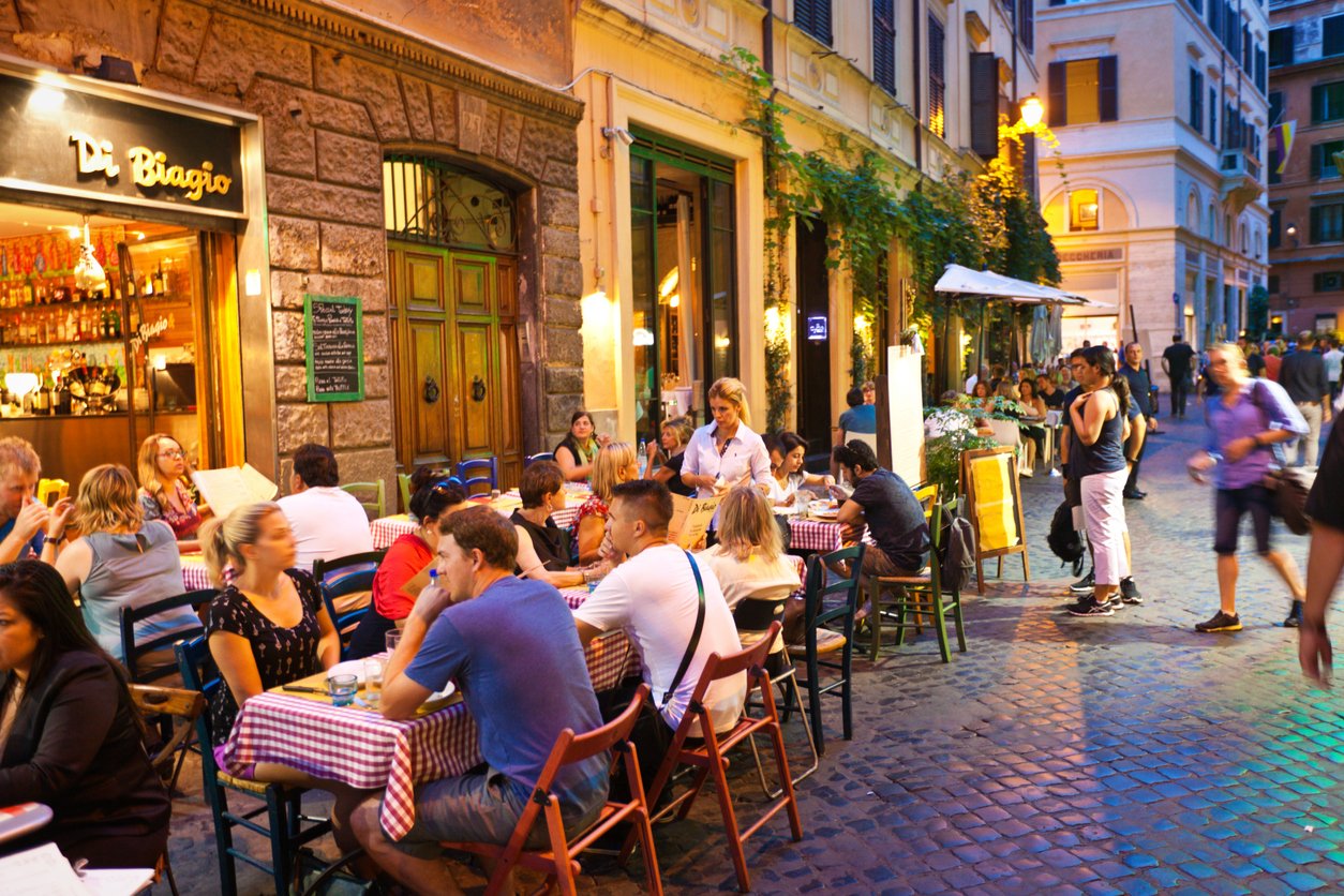 European cobblestone street, outdoor dining, sidewalk restaurants, dusk with warm lights, editorial travel photo, people present.
