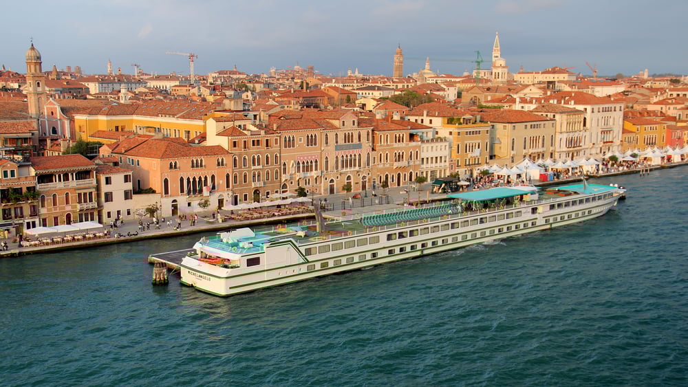 Venice waterfront, river cruise ship foreground, colorful facades and terracotta roofs, daylight clouds, editorial travel photo, no people.