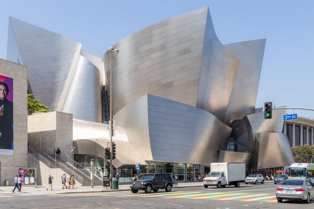 Walt Disney Concert Hall, gleaming metal curves, street intersection foreground, midday sunlight, editorial travel photo, people visible.
