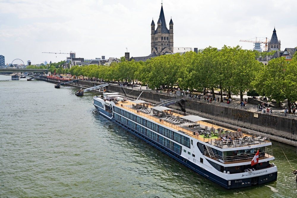 River promenade, cruise ship docked, historic buildings and twin-spired church, wide view, daylight, editorial travel photo, no people.