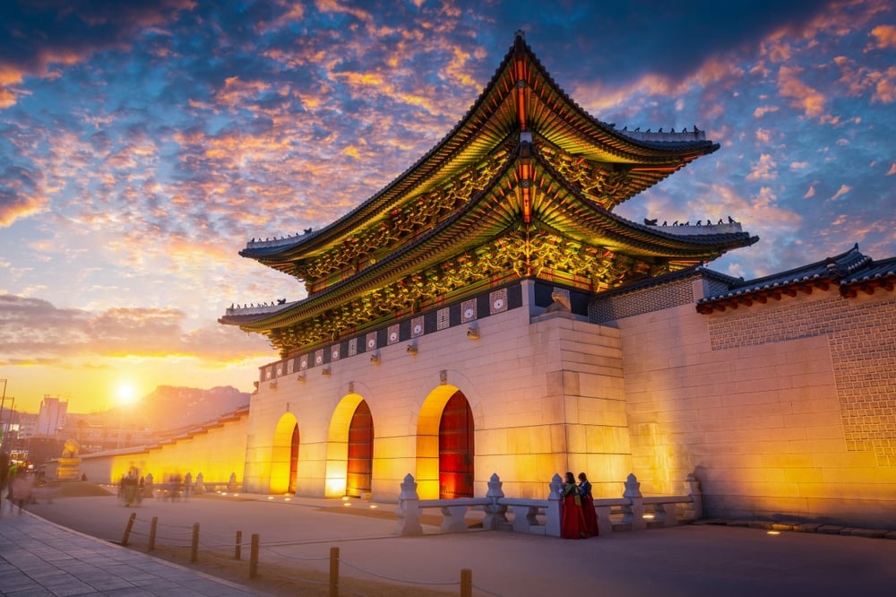 Korean palace gate, ornate wooden doors, sunset sky colors, evening glow, editorial travel photo, three people in hanbok.