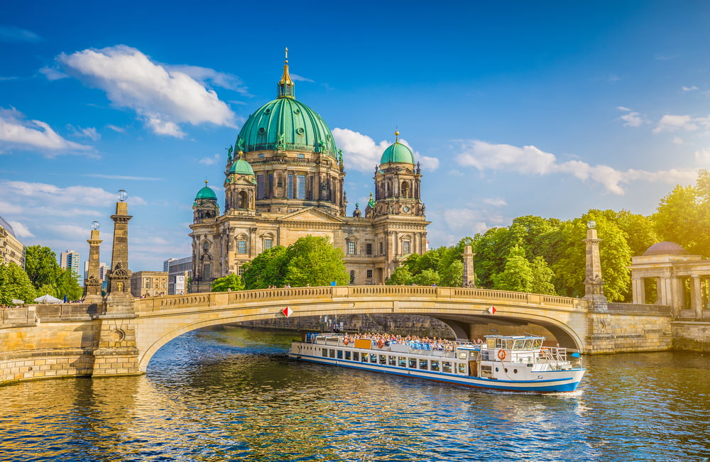 Berlin Cathedral, white river cruise boat, under stone bridge, midday sunlight, editorial travel photo, no people visible.