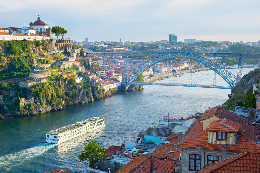 Porto riverfront, cruise boat beneath Dom Luís I Bridge, terracotta rooftops, midday light, editorial travel photo, no people.