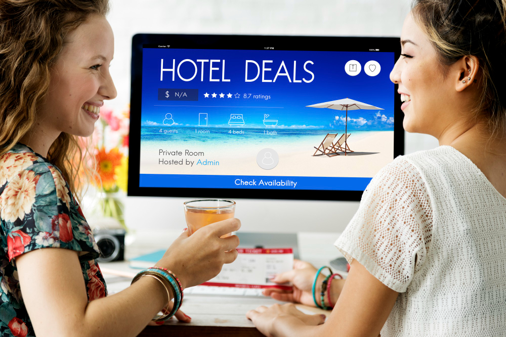 Home office, hotel deals on monitor, casual drinks in hand, daylight through window, editorial travel photo, two women present.