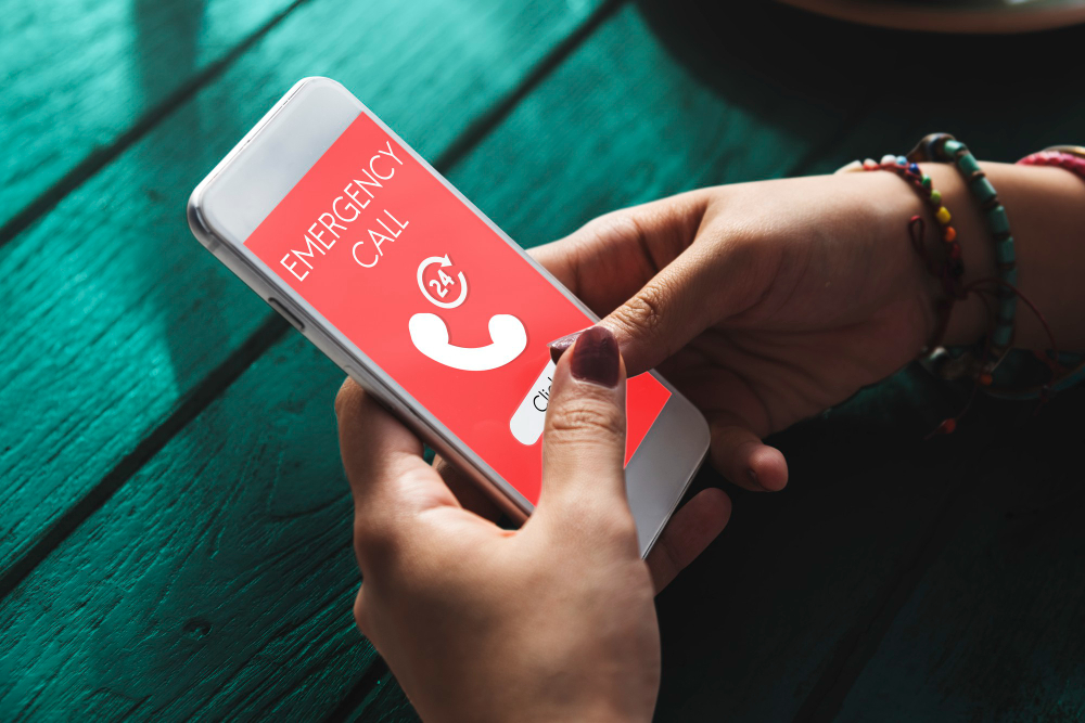 Indoor setting, smartphone with red EMERGENCY CALL screen foreground, close-up, natural light, documentary style, person visible.