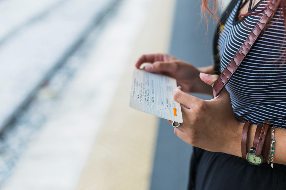 Train station platform, ticket in hand foreground, striped shirt mid-frame, daytime natural light, editorial travel photo, no faces shown.