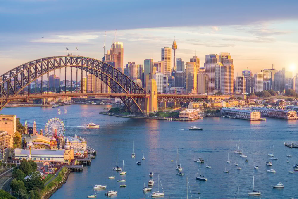 Sydney cityscape, Harbour Bridge focal point, waterfront amusement park and ferris wheel, sunset glow, editorial travel photo, no people.
