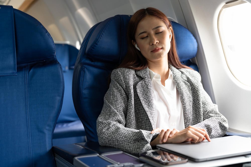 Airplane cabin, businesswoman relaxing, window seat with devices on tray table, daytime light, editorial travel photo, one person.