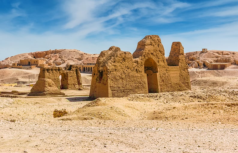 Desert ruins, mudbrick walls foreground, sandy hills backdrop, midday sun, editorial travel photo, no people.