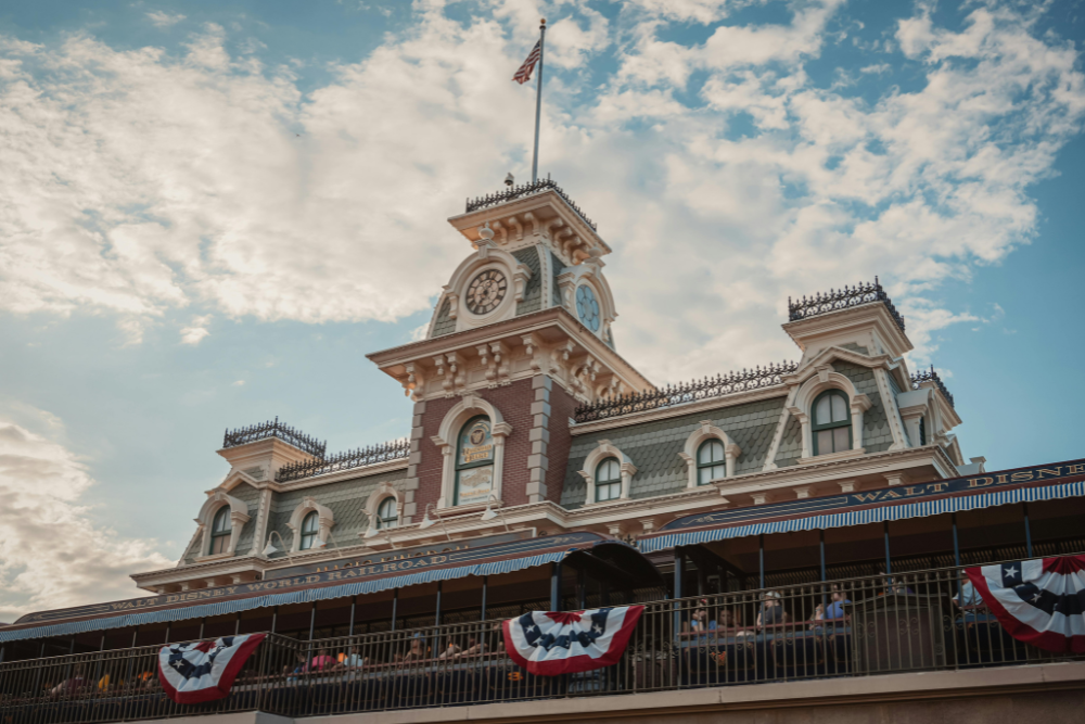 Walt Disney World Railroad Main Street Station, clock tower, patriotic bunting, partly cloudy day, editorial travel photo, no people.