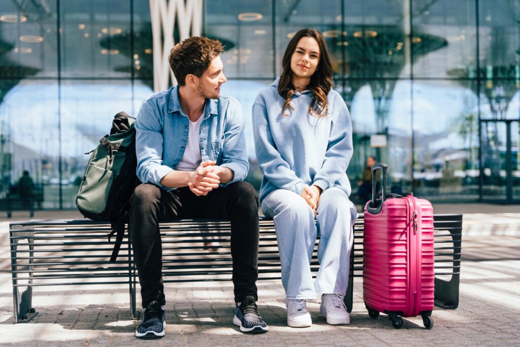 Modern building exterior, pink suitcase focal point, bench foreground, daytime natural light, editorial travel photo, two people.