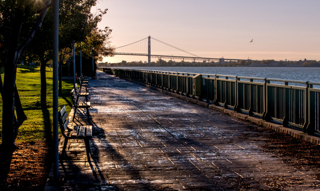 Riverside walkway, empty benches, distant suspension bridge centered, sunrise glow, editorial travel photo, no people.