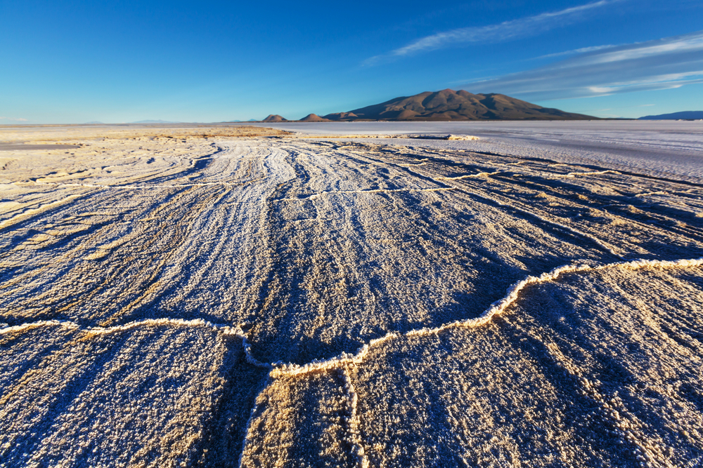 Salt flats, cracked patterns foreground, receding lines to distant mountains, midday clear blue sky, editorial travel photo, no people.