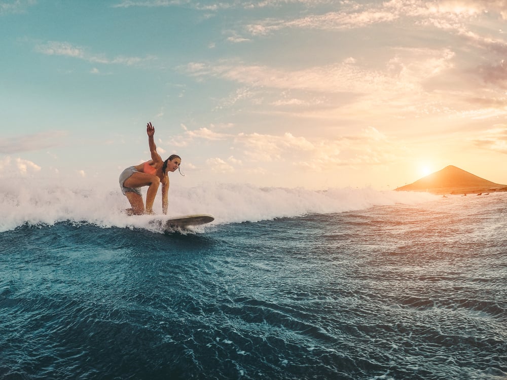 Ocean at sunset, surfer on a wave, island silhouette background, colorful sky glow, editorial travel photo, one person.