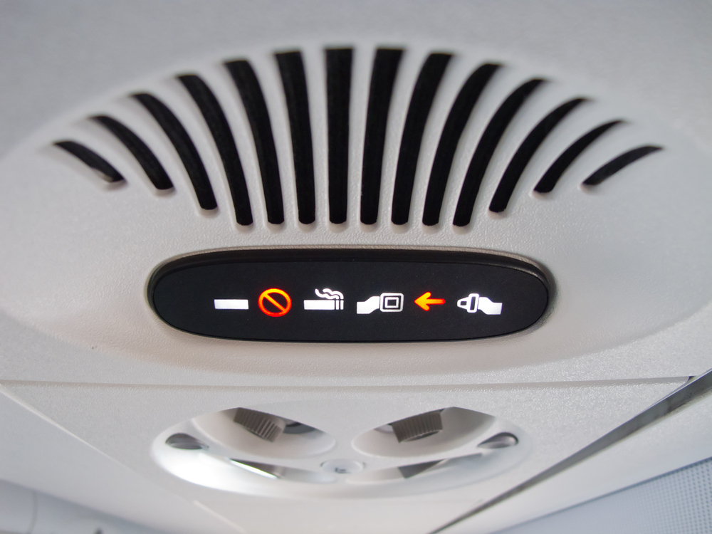 Airplane cabin overhead panel, illuminated signs detail, tight crop, artificial lighting, editorial travel photo, no people.