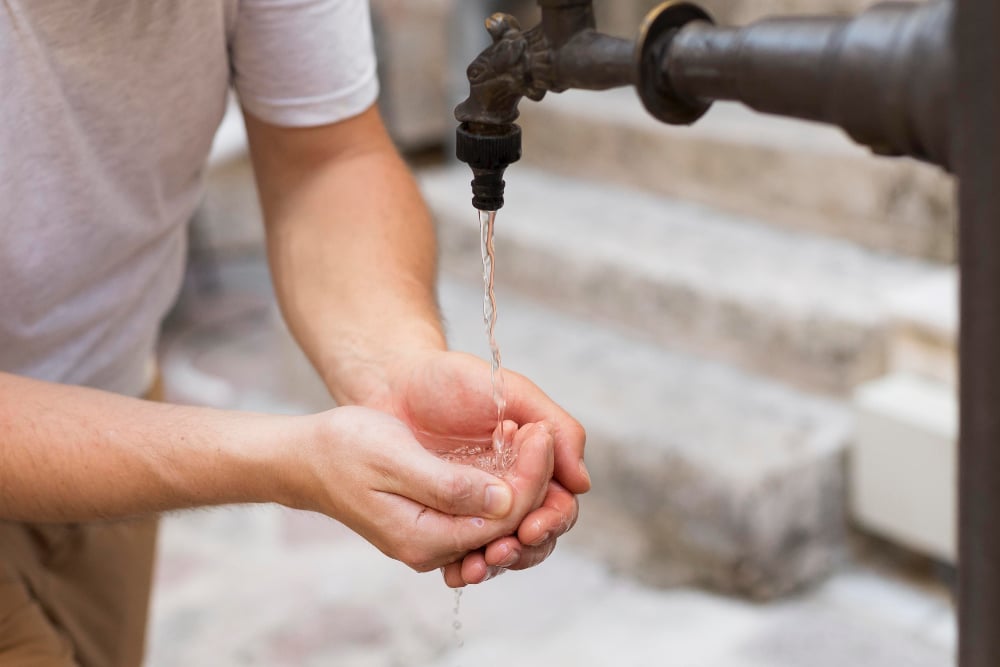 Outdoor faucet, cupped hands catching water, mid-frame composition, daylight, editorial travel photo, person present.