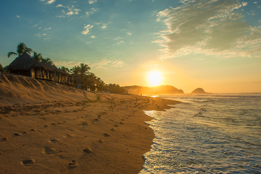 Sandy beach, sunset sky, palm trees left, golden hour glow, editorial travel photo, no people.