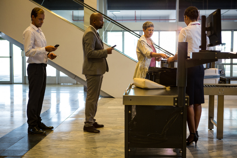 Airport security checkpoint, traveler placing items in tray, line formation, bright lighting, editorial travel photo, people present.
