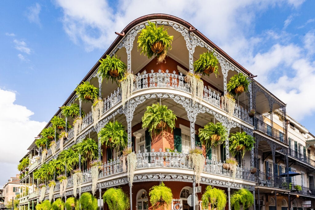 Three-story building with ornate iron railings, numerous hanging plants, and a red brick facade under a blue sky.