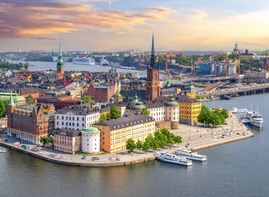 Aerial view of Stockholm's old town with historical buildings, waterfront, and cruise ships under a colorful sky.