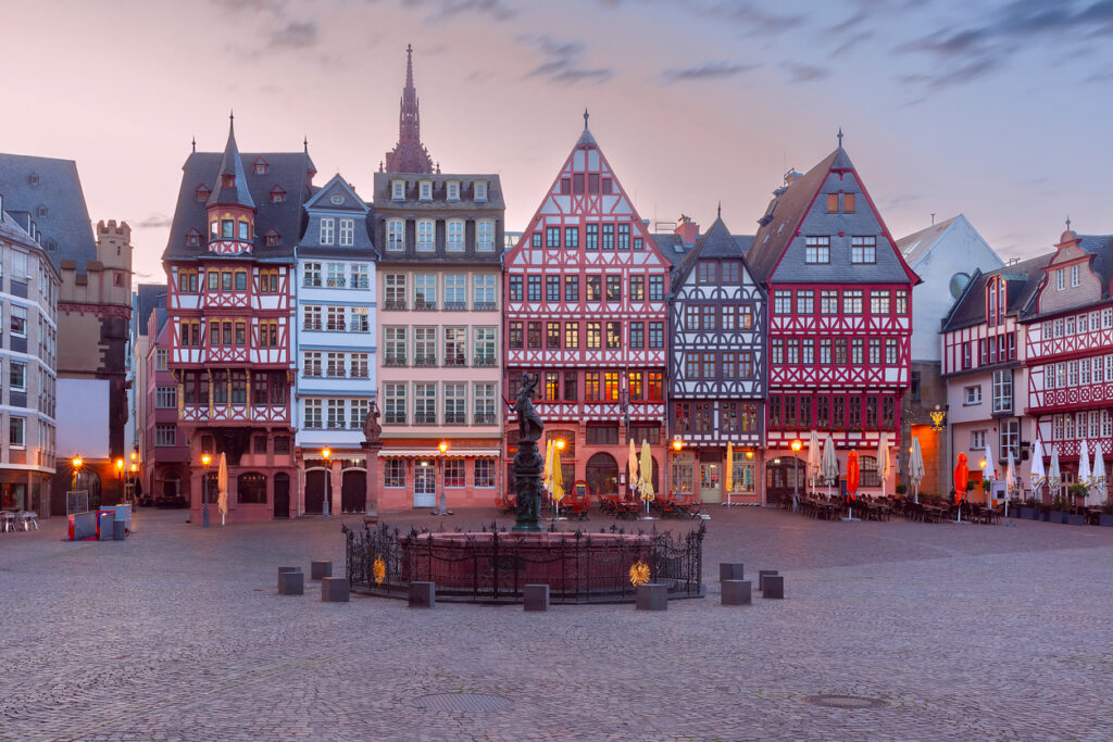 Historic square with half-timbered buildings, evening lighting, cobblestone ground, and fenced statue in the center.