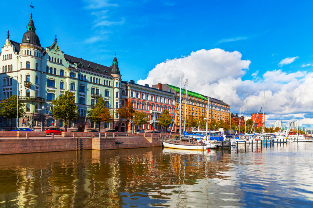 Helsinki waterfront, sailboats docked, buildings and trees reflected in still water, daytime light, editorial travel photo, no people.