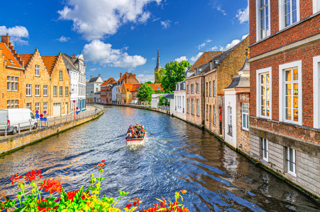 Boat with passengers on a canal, surrounded by brick buildings and colorful flowers, under a blue sky.