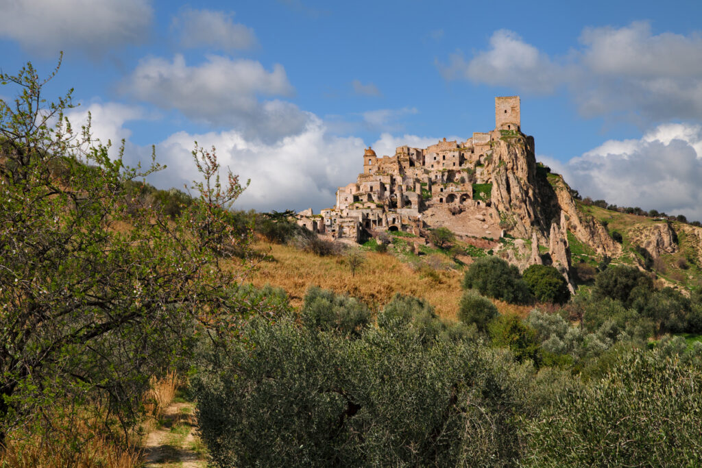 Hilltop village with stone buildings and a tower, surrounded by trees and greenery under a blue sky.