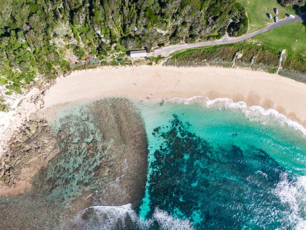 Aerial view of beachfront with clear turquoise water, sandy shore, and adjacent greenery and road.