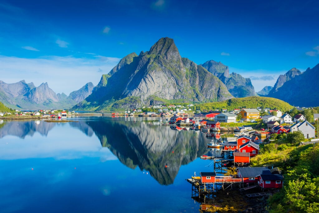 Norwegian village, red waterfront houses, nestled by blue fjord and mountains, midday sunlight, editorial travel photo, no people.