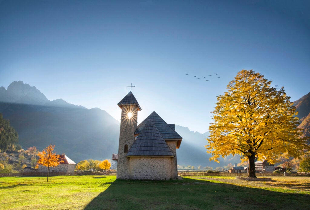 Stone church, yellow autumn tree, bordered by field and mountains, clear daylight, editorial travel photo, no people.