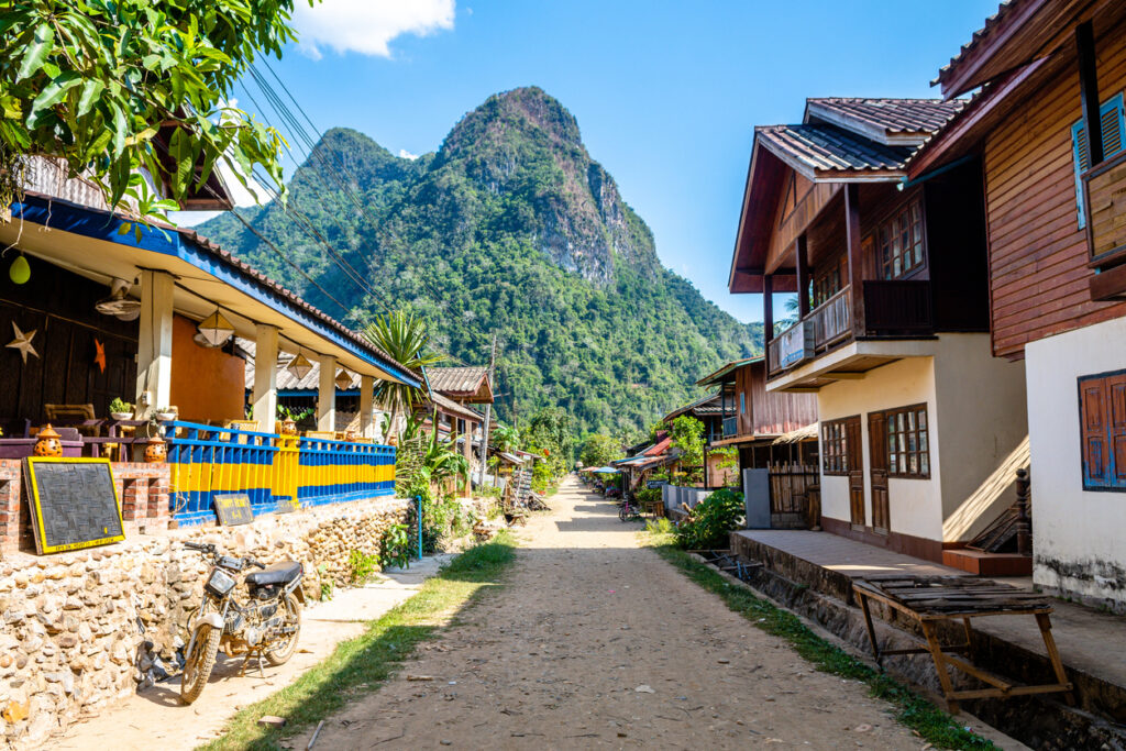 Dirt road lined with wooden buildings and greenery, leading to a large, lush mountain under a blue sky.
