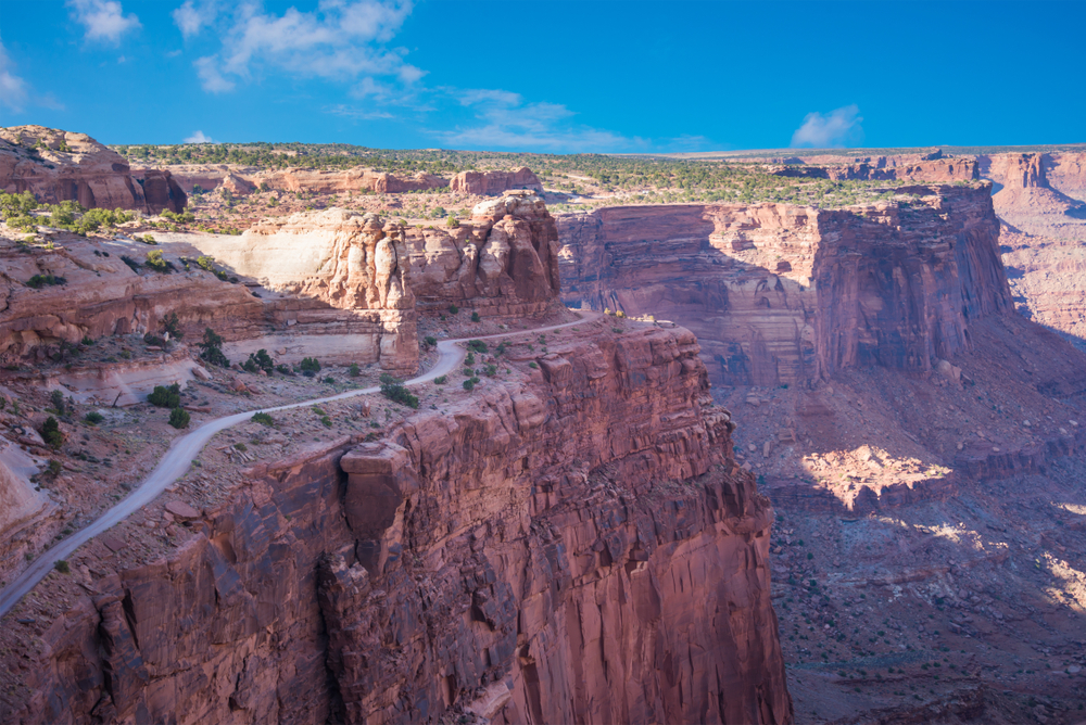 A winding dirt road along a steep red rock cliff under a blue sky with light clouds.