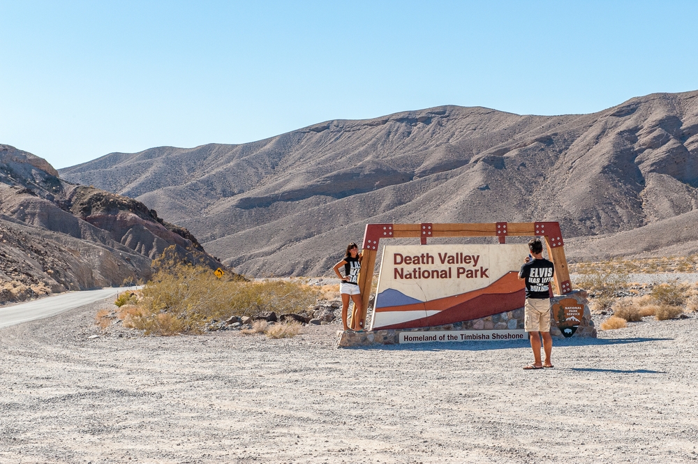 Two people stand by the Death Valley National Park sign, mountainous desert landscape in the background.