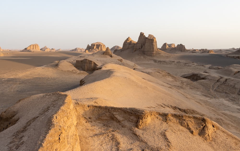 A desert landscape with large rock formations under a clear sky, featuring sandy dunes and sparse vegetation.