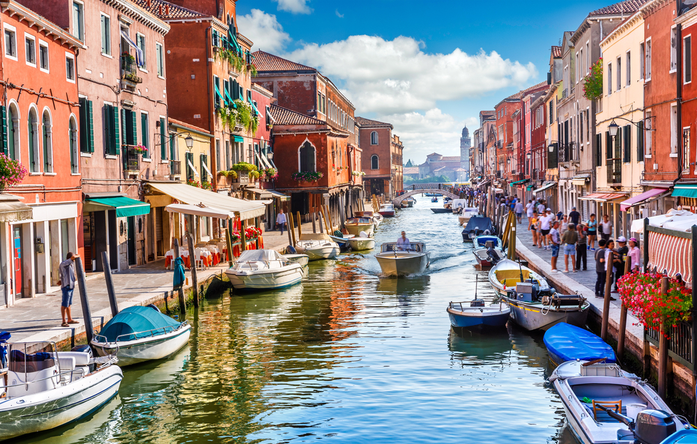 Narrow canal with boats, colorful buildings on both sides, and people walking along the sidewalks.