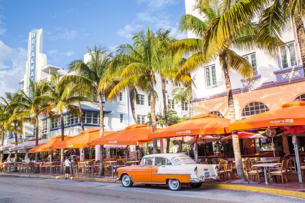 Ocean Drive, vintage orange and white car foreground, outdoor cafés, midday sunlight, editorial travel photo, no people.